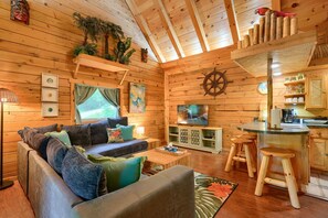 Ein gemütliches Blockhaus-Interieur mit Holzbalkendecke, einer Couch, einem Fernseher, einer Küchenzeile und einem Fenster mit Vorhängen.