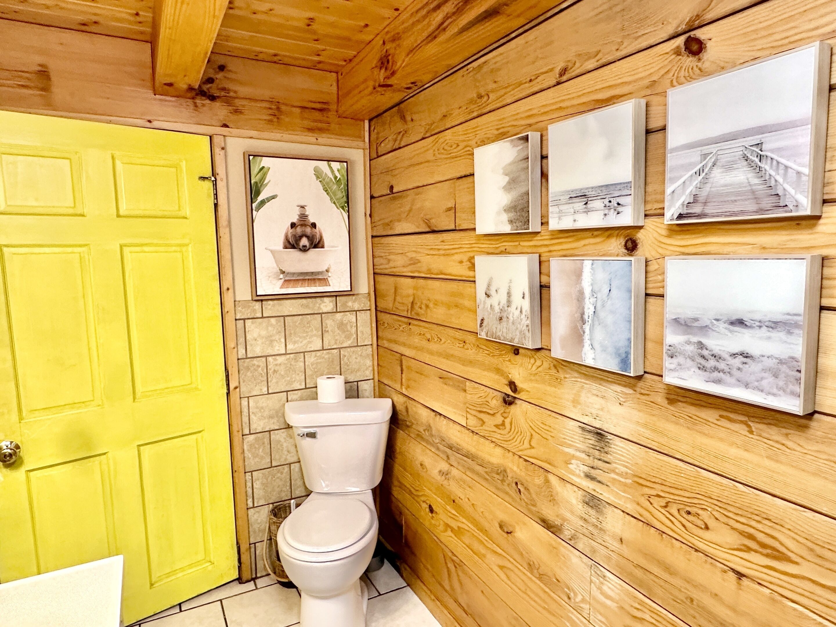 Un baño con paneles de madera, una puerta amarilla, un inodoro y varias fotografías enmarcadas en la pared.
