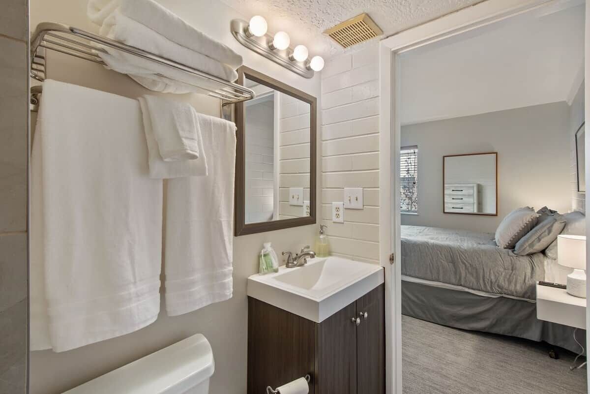 A bathroom with a sink, mirror, towel rack, and a bed visible in the adjacent room.