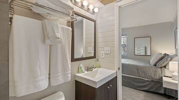 A bathroom with a sink, mirror, towel rack, and a bed visible in the adjacent room.