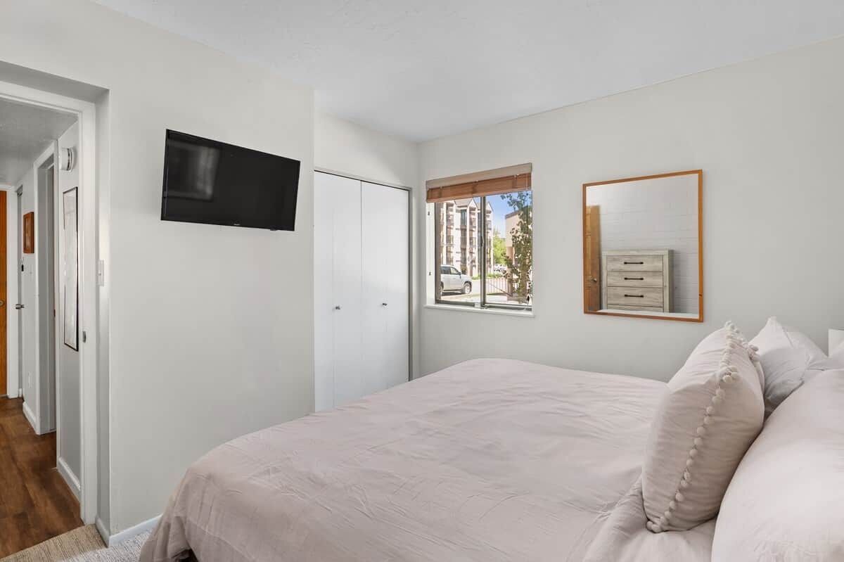 Une chambre à coucher avec un lit, un téléviseur fixé au mur, une fenêtre donnant sur des immeubles et une porte.