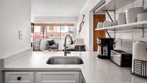 A kitchen with a sink, coffee maker, toaster, and shelves with dishes. - Prospector Place - 2(King)Bed 2Bath - Easy to ski! (Park City)