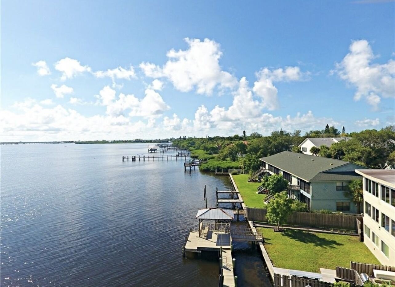 A waterfront property with a dock extending into a calm body of water, surrounded by greenery and residential buildings.