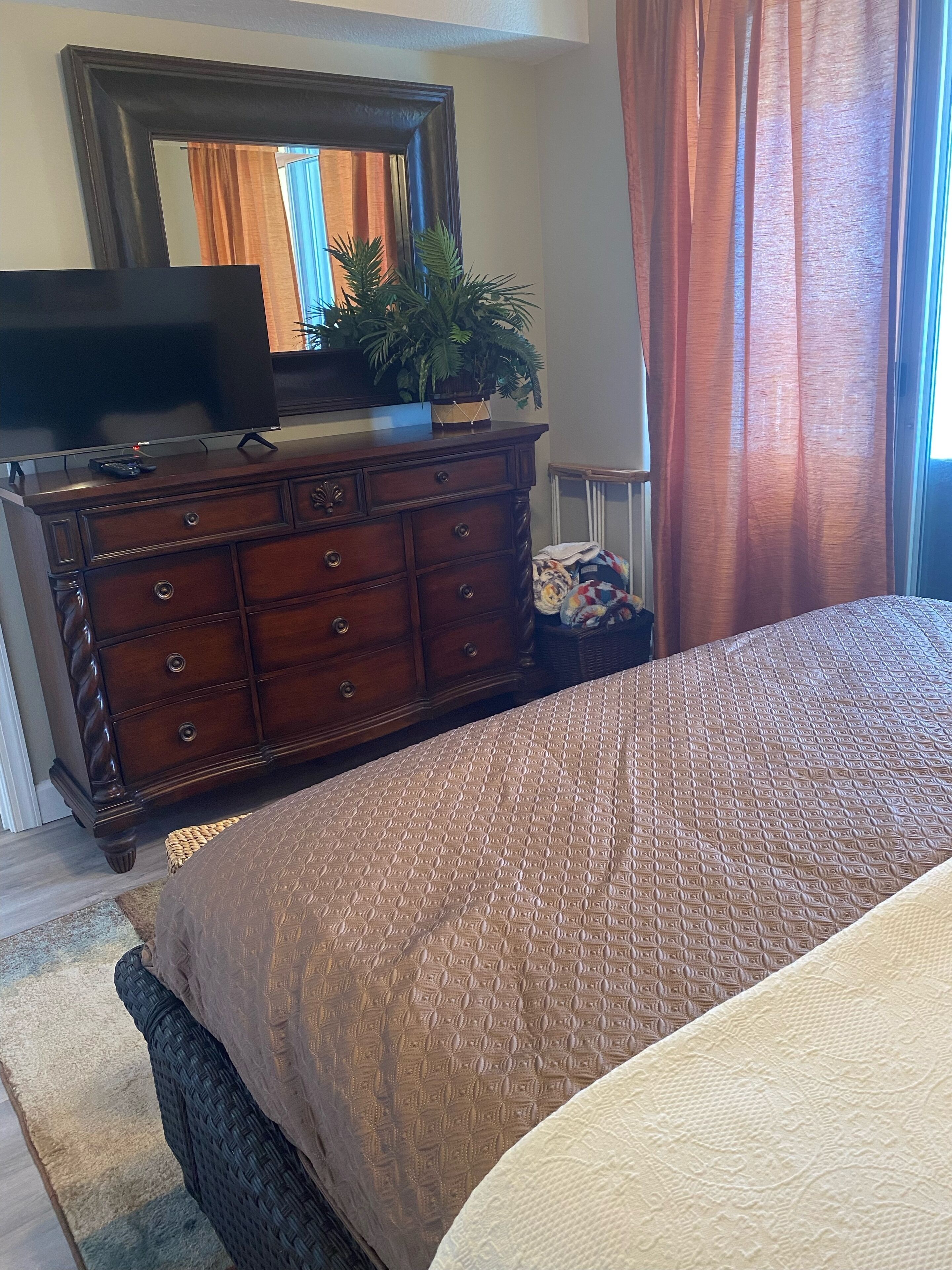 A bedroom with a large bed, a dresser with a mirror, a television on the dresser, and a window with curtains.