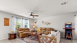 Ein Wohnzimmer mit braunem Ledersofa, einem Couchtisch, einem gemusterten Teppich, einem Deckenventilator und einer Spielkonsole.