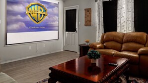 Ein Wohnzimmer mit einem großen Bildschirm, der das Warner Bros. Pictures Logo zeigt, einem braunen Ledersofa, einem Holztisch und einem kleinen Beistelltisch mit einer Vase.