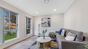 A modern living room with a grey sofa, a round coffee table, and a floor lamp. There is a large window providing a view of the outside.