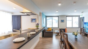 Una cuina moderna amb una illa central, zona de menjador amb cadires de fusta i un sofà amb coixí groc.