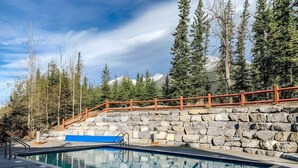 A swimming pool with a stone wall and wooden railing, surrounded by trees and a clear sky. - Top-Floor Condo with stunning mountain views! (Canmore)