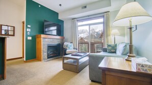 A living room with a fireplace, a television, a sofa, and a window with a view of trees. - Top-Floor Condo with stunning mountain views! (Canmore)
