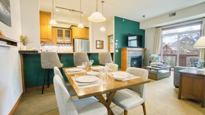 A dining area with a wooden table set for four, surrounded by four chairs. There is a kitchen area with wooden cabinets and a built-in oven. A fireplace is visible in the background, and a television is mounted above it. - Top-Floor Condo with stunning mountain views! (Canmore)