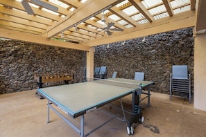 Mesa de ping pong al aire libre con pared de piedra de fondo, ventiladores de techo y estructura de pérgola.