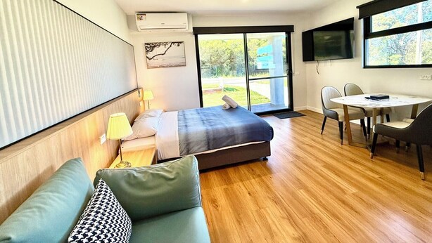 Una habitación de hotel moderna con cama, sofá, mesa de comedor con sillas, televisor y un amplio ventanal con vistas al exterior.