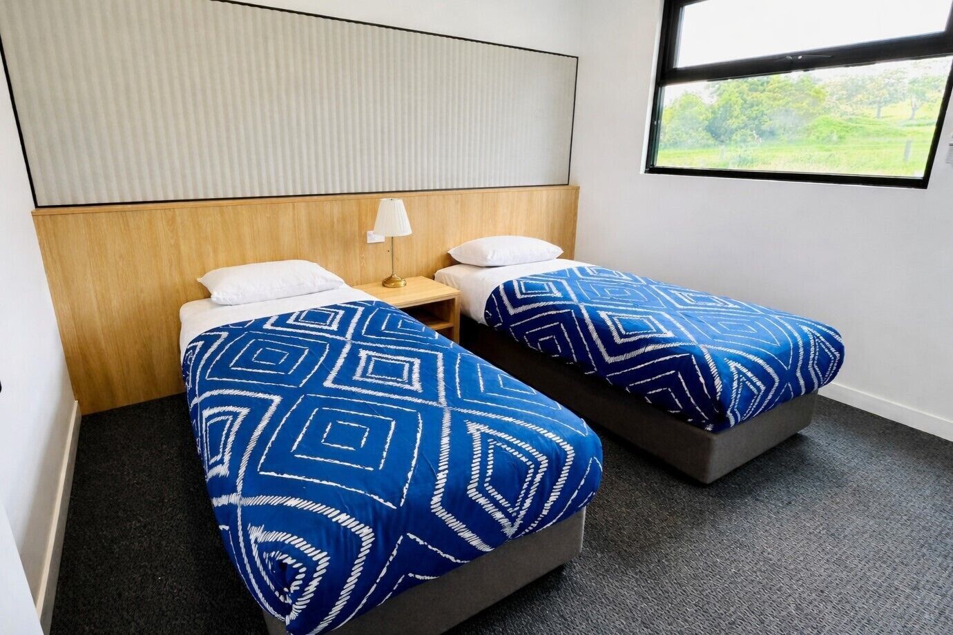 Quarto de hotel com duas camas de solteiro, cabeceira de madeira, mesa de cabeceira com abajur e janela com vista para o verde.