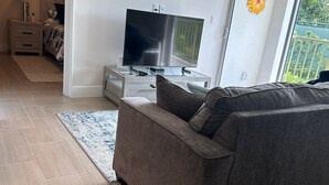 A modern living room with a grey sofa, a flat-screen TV on a wooden stand, and a large sliding glass door leading to a balcony.