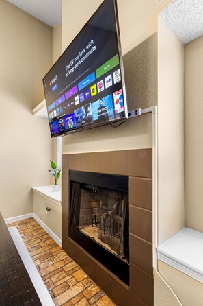 A modern living room with a flat-screen TV mounted above a fireplace, a small plant on a shelf, and a stone-patterned floor. - Westside Home: 2 King, 2 Queen, BBQ Oasis (El Paso)