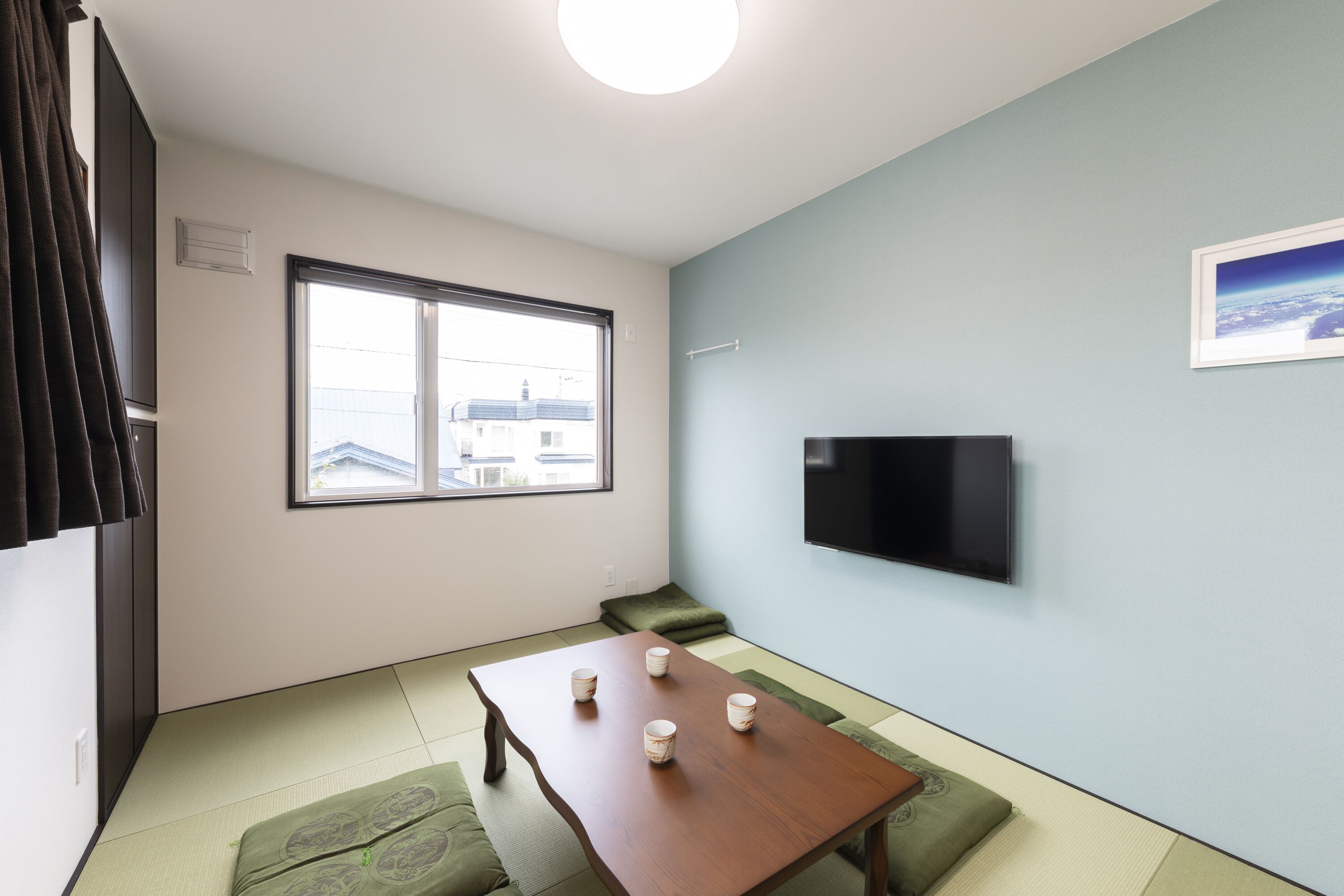 A room with tatami flooring, a low table with four cups, a green cushion, a TV on the wall, and a window with a view of buildings.