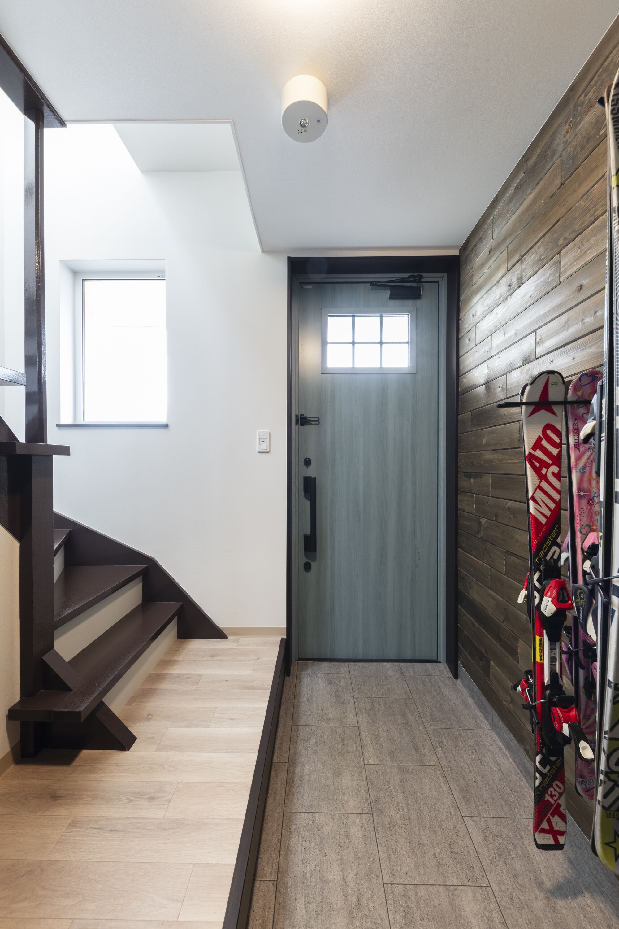 A modern interior with a wooden staircase, a door with a window, and skis leaning against a wall.