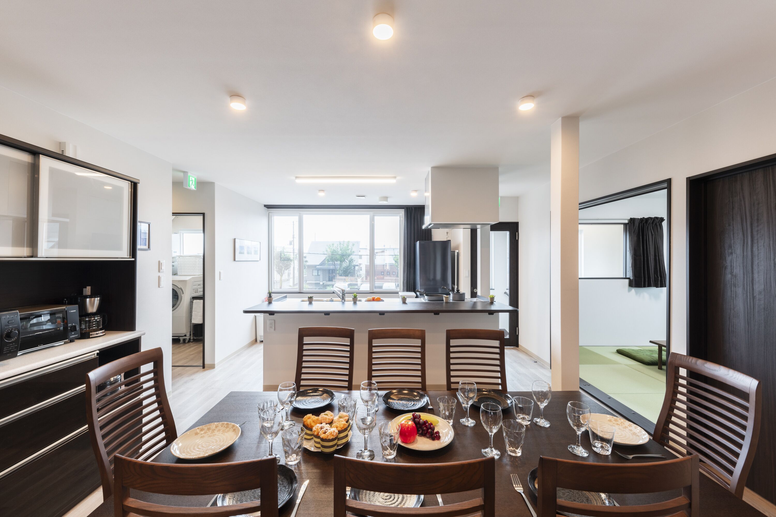 A modern dining area with a large table set for six, surrounded by wooden chairs. A kitchen area is visible in the background with a microwave and other appliances.