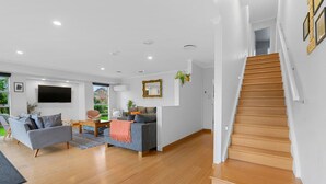 Un salón amplio con escalera de madera, un sofá gris y un televisor de pantalla plana montado en la pared.