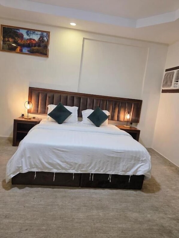 Een net opgemaakt bed met een houten hoofdbord, twee nachtkastjes met lampen, en een schilderij aan de muur.