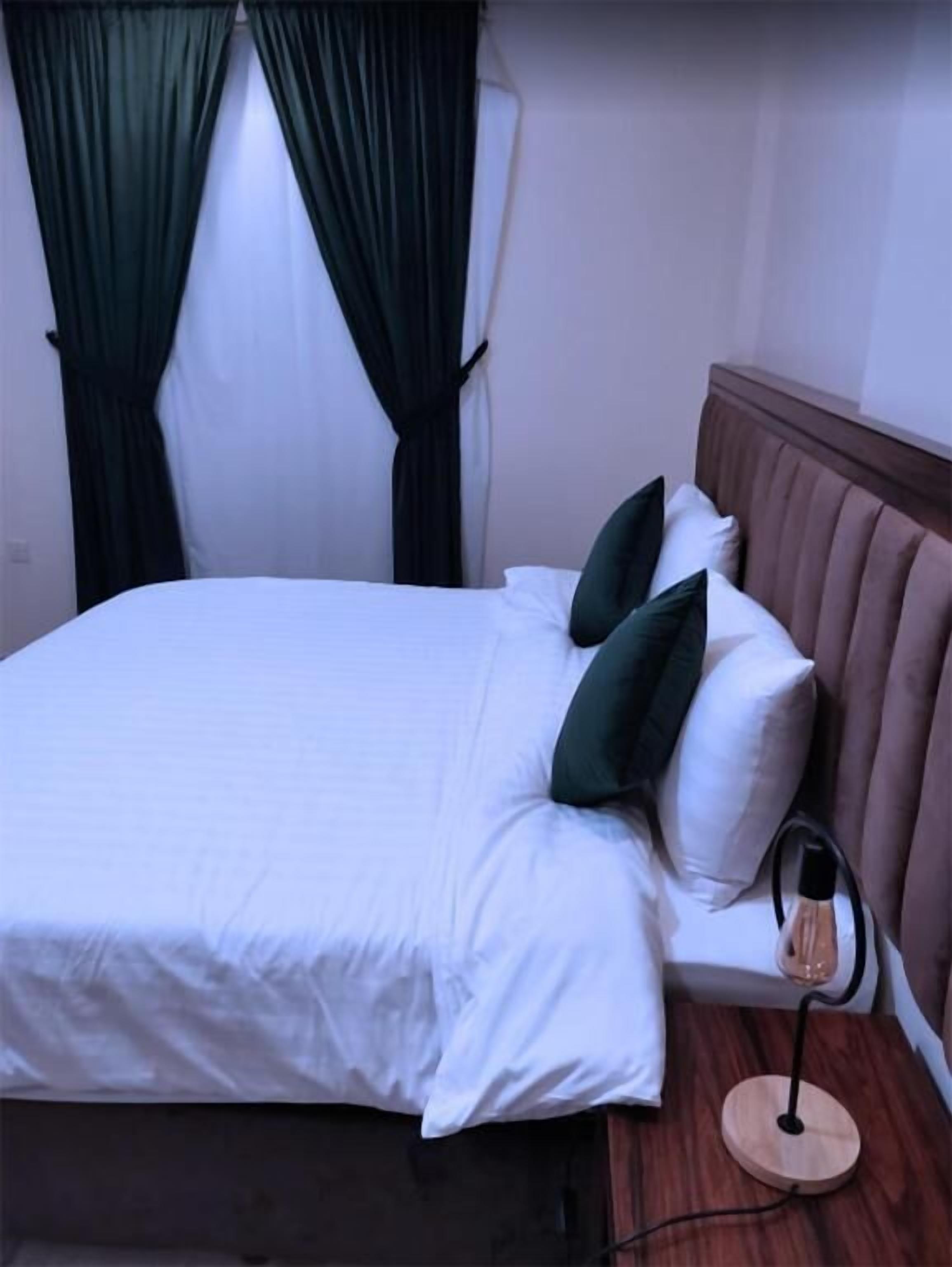 Ein ordentlich bezogenes Bett mit weißen Leinen und zwei grünen Kissen, ein Nachttisch mit Lampe und dunkle Vorhänge am Fenster.