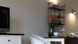 A modern kitchen with a white dining table, a white chair, a TV, and a shelf with glassware and flowers. - Loft - Neu ab 15.01.2024 - Cityloft- Modernes Appartement für 2 Personen in Zentraler Lage (Sylt)