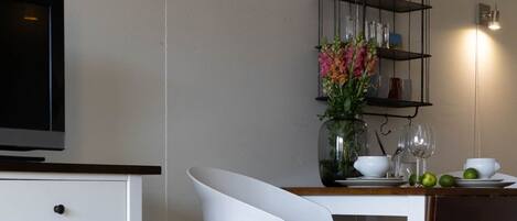 Una cocina moderna con una mesa de comedor blanca, una silla blanca, un televisor y una estantería con cristalería y flores.