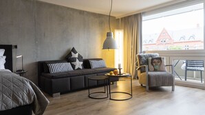 A modern hotel room with a sofa, a round coffee table, a chair, and a large window offering a view of buildings. - Loft - Neu ab 15.01.2024 - Cityloft- Modernes Appartement für 2 Personen in Zentraler Lage (Sylt)