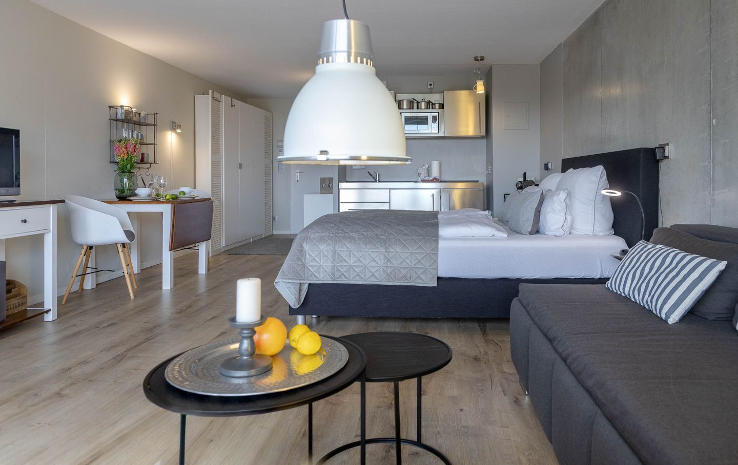 A modern bedroom with a large bed, a bedside table, a desk, and a kitchenette in the background.