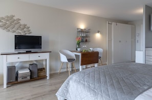 Quarto com cama, escrivaninha com televisão, cadeira, guarda-roupa e prateleira com objetos decorativos.