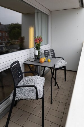 Uma varanda com mesa e cadeiras, um vaso com flores e uma janela com cortinas.