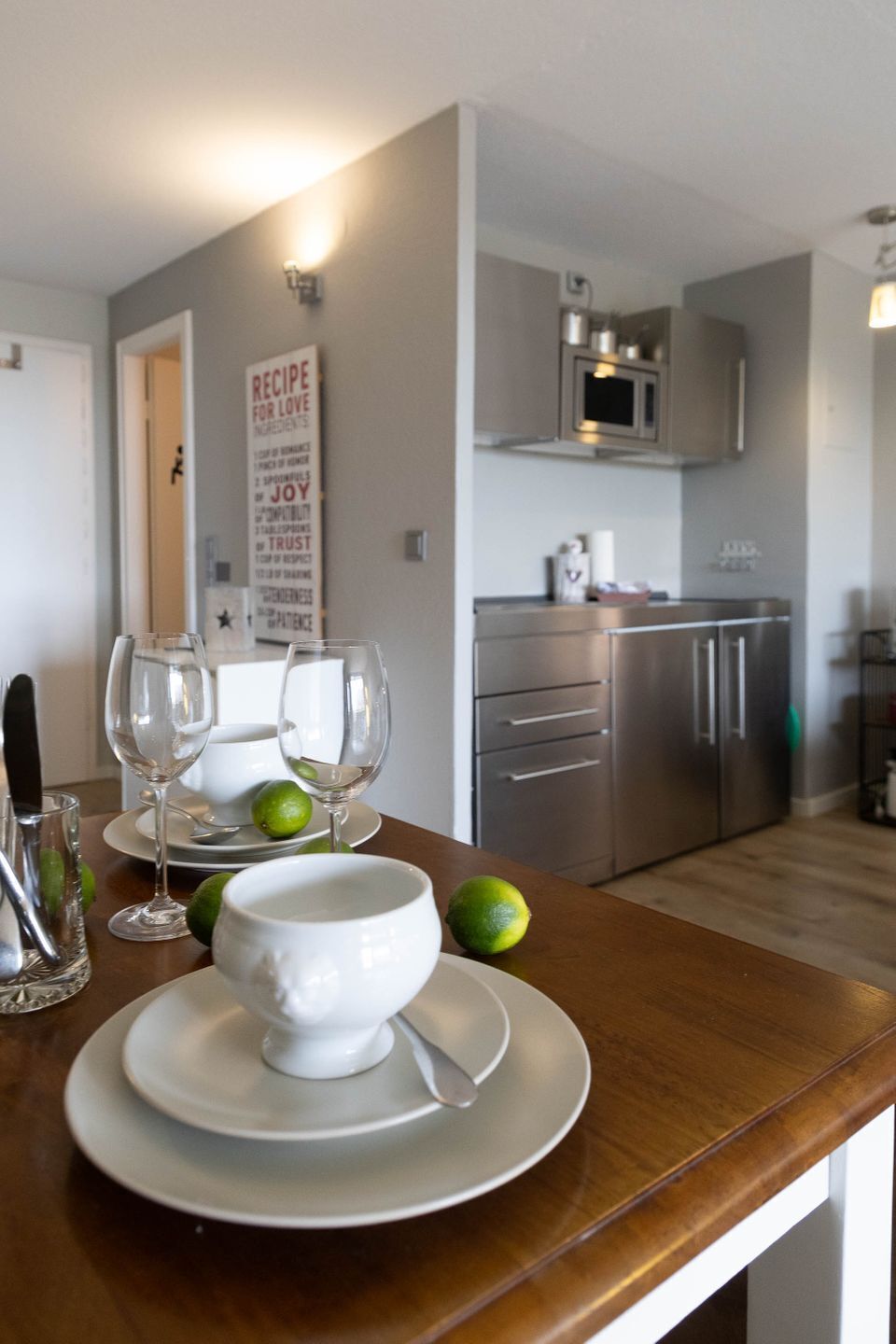 A kitchen with stainless steel appliances, a white wall with a recipe sign, and a dining area with a white bowl and glasses on a wooden table.