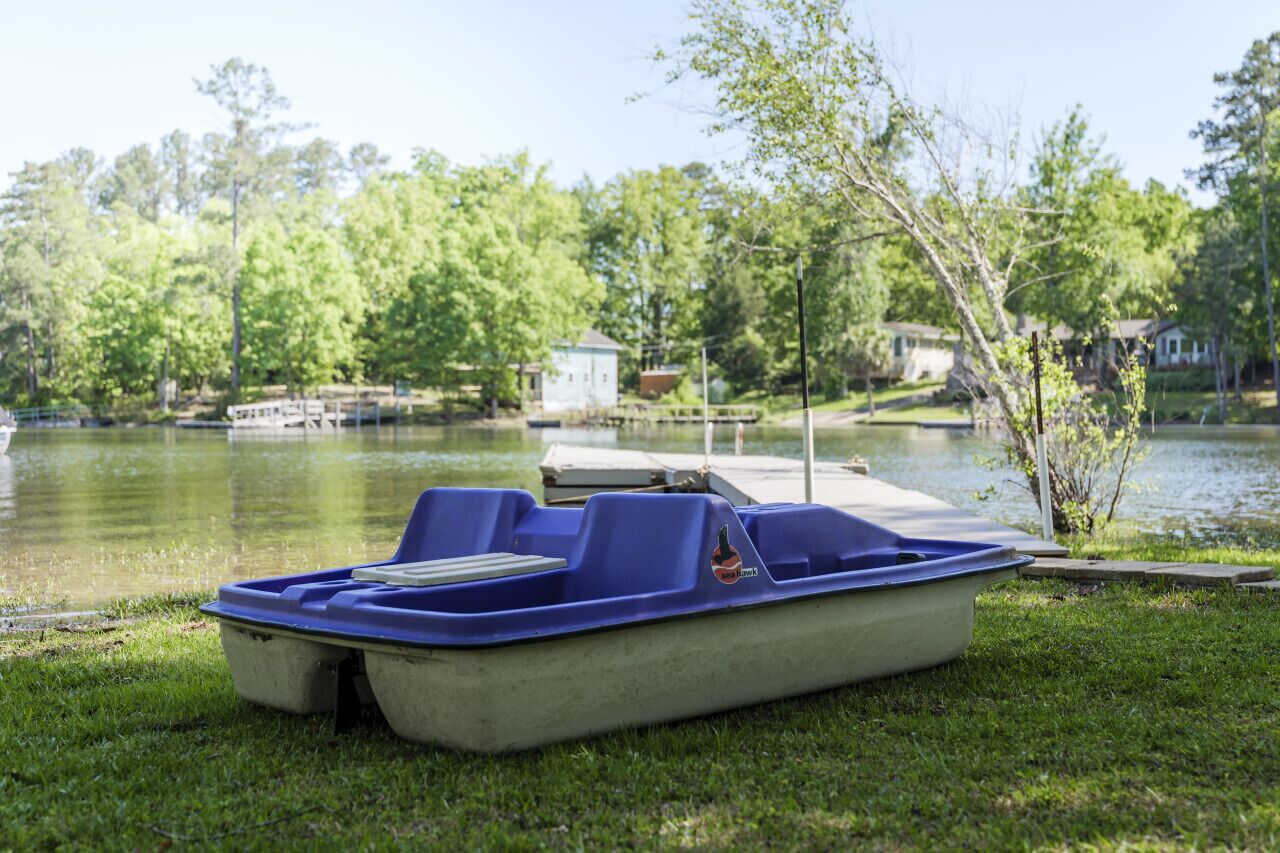 A blue pontoon boat on a grassy area near a lake with a dock and a small building in the background.