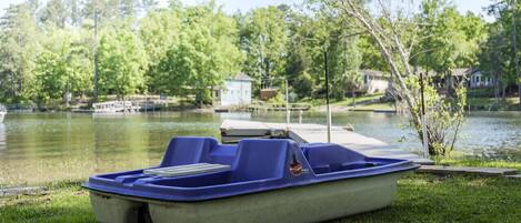 A blue pontoon boat on a grassy area near a lake with a dock and a small building in the background.