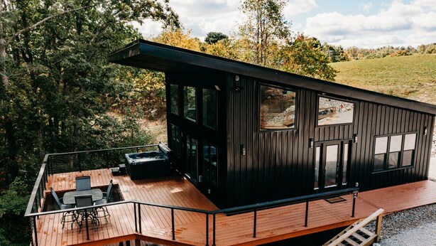 Een moderne cabin met een houten terras, een bubbelbad en een groot raam met uitzicht op een bebost gebied.