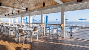 Une terrasse extérieure avec des tables et des chaises blanches, des parasols bleus et une vue sur l’océan.