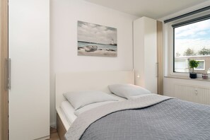 A bedroom with a bed, a painting of a lighthouse, a window with a view, and a radiator.