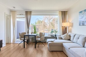A modern living room with a beige sofa, a dining table with chairs, a small table with a lamp, and a view of trees and buildings outside.