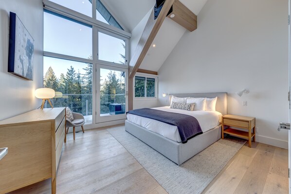 A modern bedroom with a large bed, a desk, and a chair, featuring large windows and a view of trees.