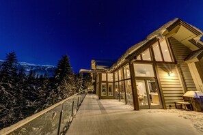 A modern building with large windows and a covered outdoor area, surrounded by snow-covered trees and mountains in the background.