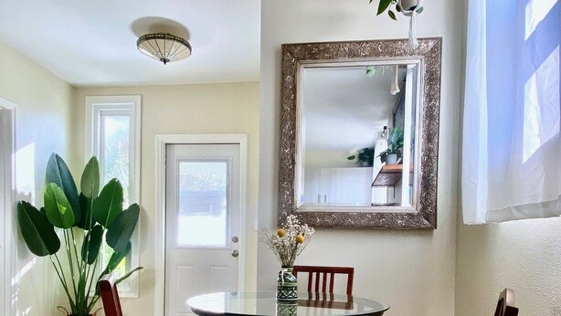 A dining area with a round glass table, four wooden chairs with cushioned seats, a decorative mirror, and a potted plant.
