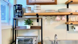 A kitchen with a toaster, microwave, coffee maker, and a sink. There is a framed picture on the wall and shelves with plates and glasses.