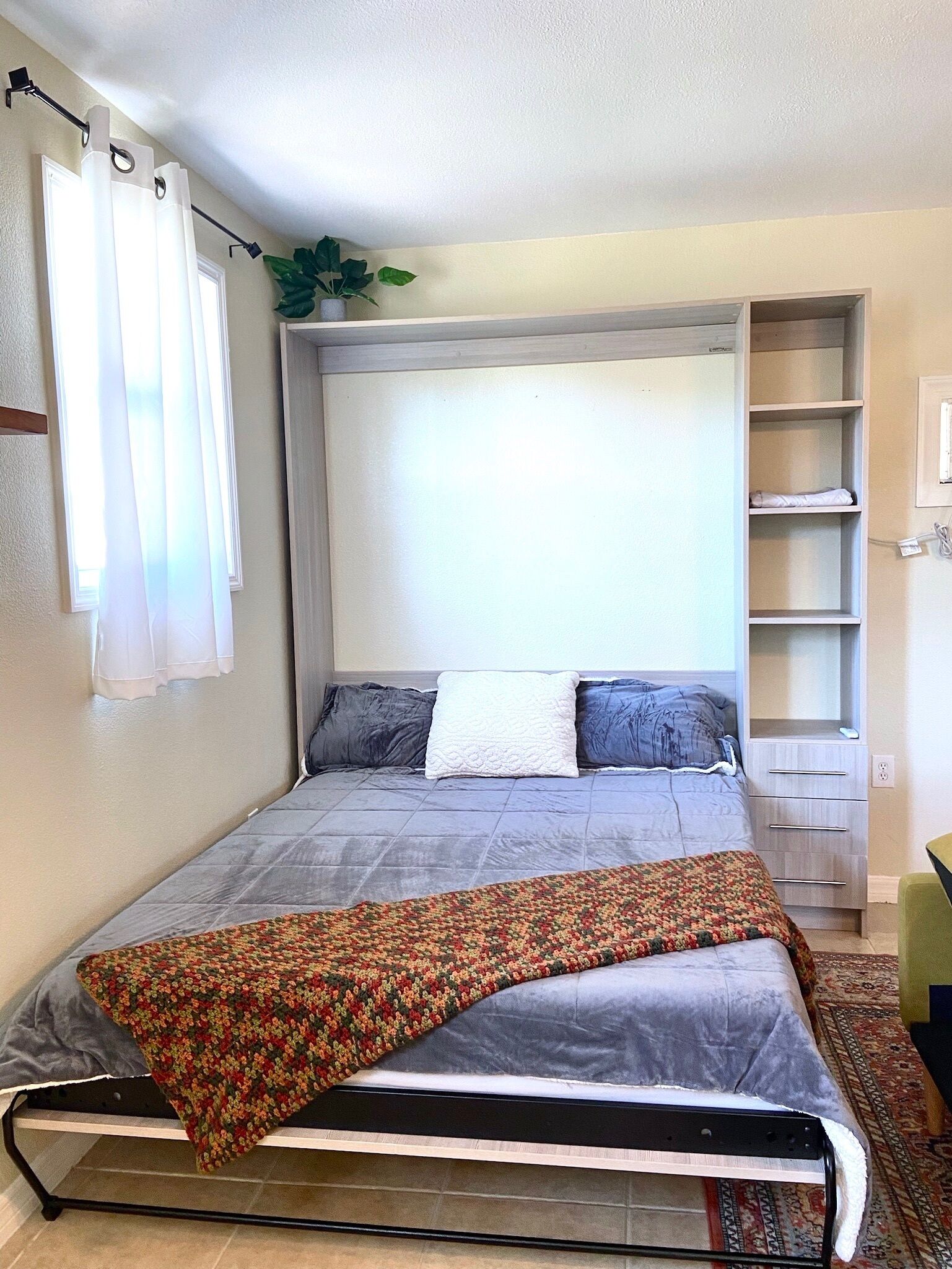 A bedroom with a bed, a built-in wardrobe, a plant, and a window with curtains.