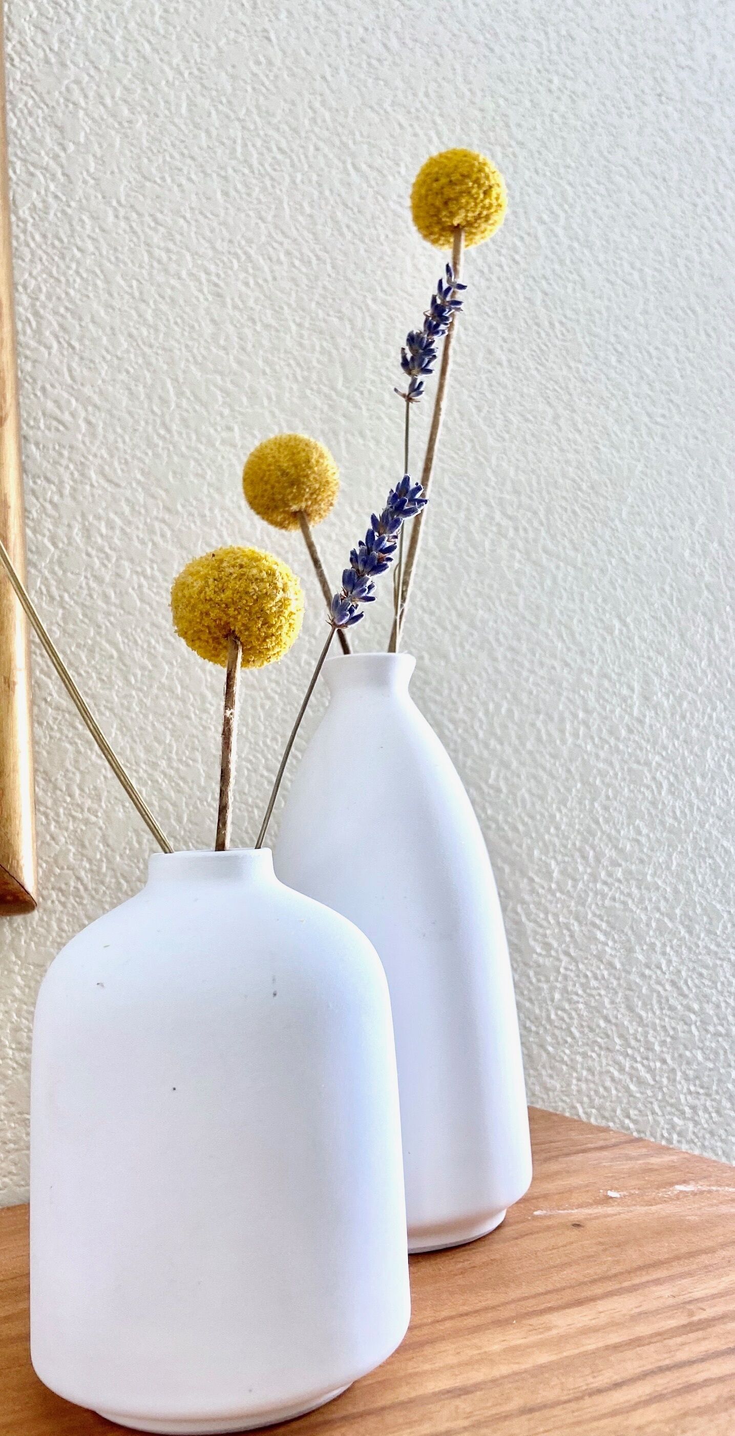 Zwei weiße Vasen mit gelben, kugelförmigen Verzierungen und violetten Blumen auf einer Holzoberfläche vor einer strukturierten Wand.