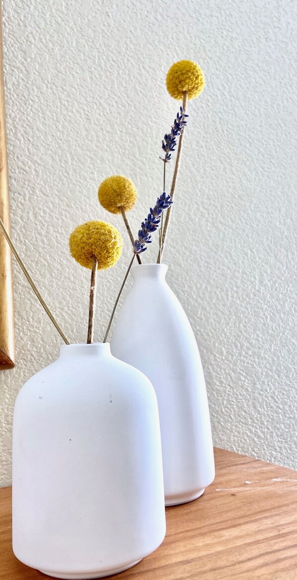 Dos jarrones blancos con decoraciones esféricas amarillas y flores moradas sobre una superficie de madera, frente a una pared con textura.