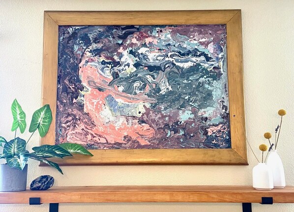 A framed abstract painting on a wall, a wooden shelf with a potted plant and a vase with dried flowers.