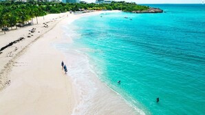 Eine Vogelperspektive zeigt einen Strand mit klarem türkisblauem Wasser, einer Sandküste und einigen Personen, die am Strand entlanggehen.