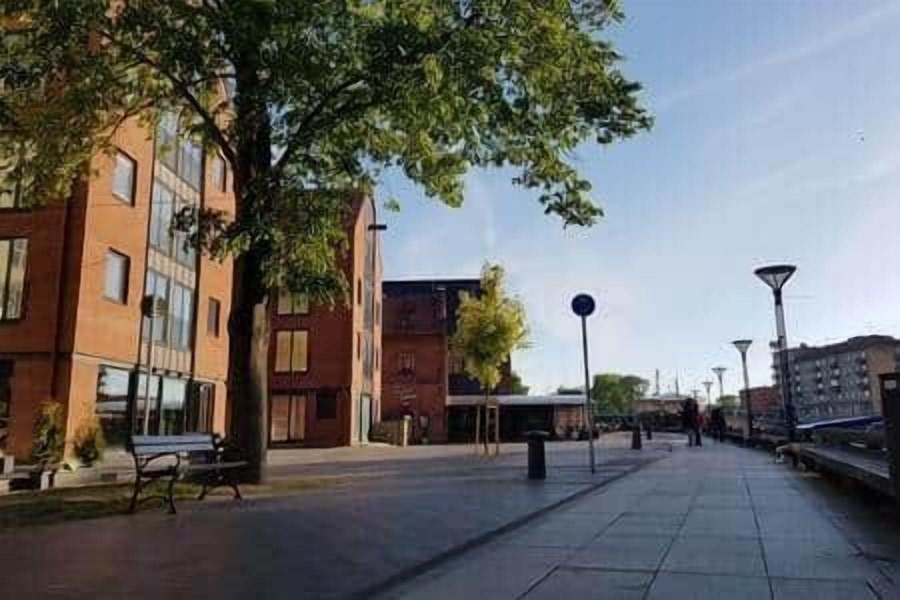 A paved walkway with benches, a trash bin, and modern buildings with large windows.