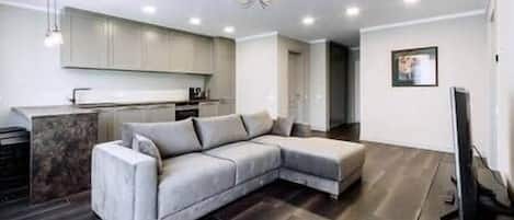 A modern living room with a sectional sofa, a flat-screen TV, and a kitchen in the background.
